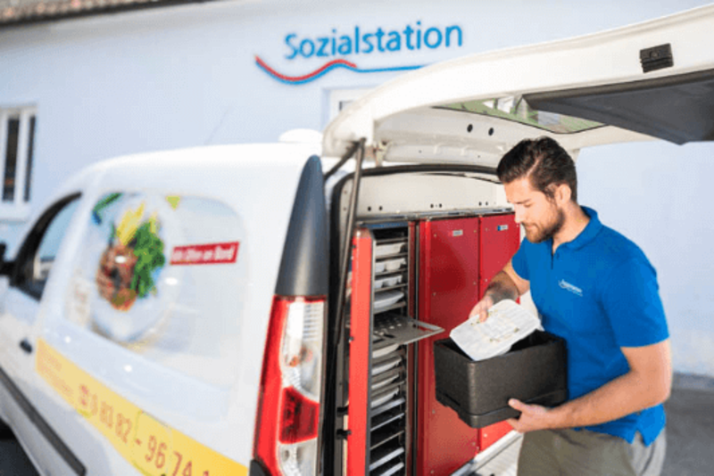 Ein Mann stellt Mahlzeiten in einen Lieferwagen vor einem Gebäude mit der Aufschrift „Sozialstation“ ab.