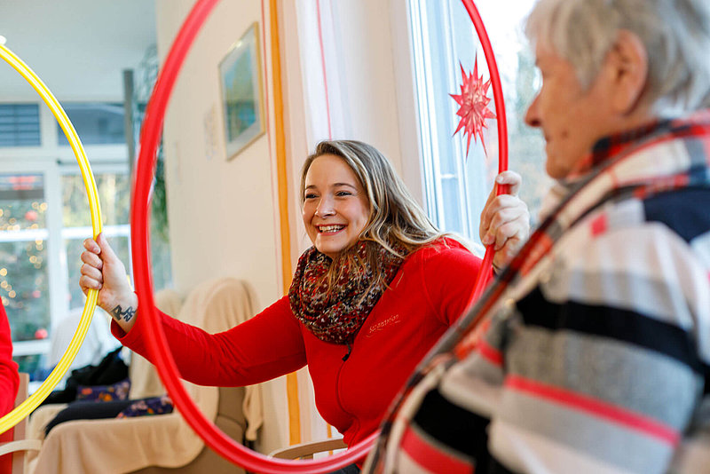 Zwei Frauen, eine in Rot lächelnd, halten bunte Reifen in einem hellen Raum mit einem Fenster und festlicher Dekoration.